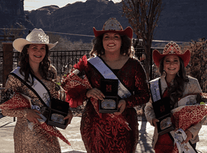 Three cowgirls in sparkly dresses and cowboy hats holding awards and flowers at an outdoor event.